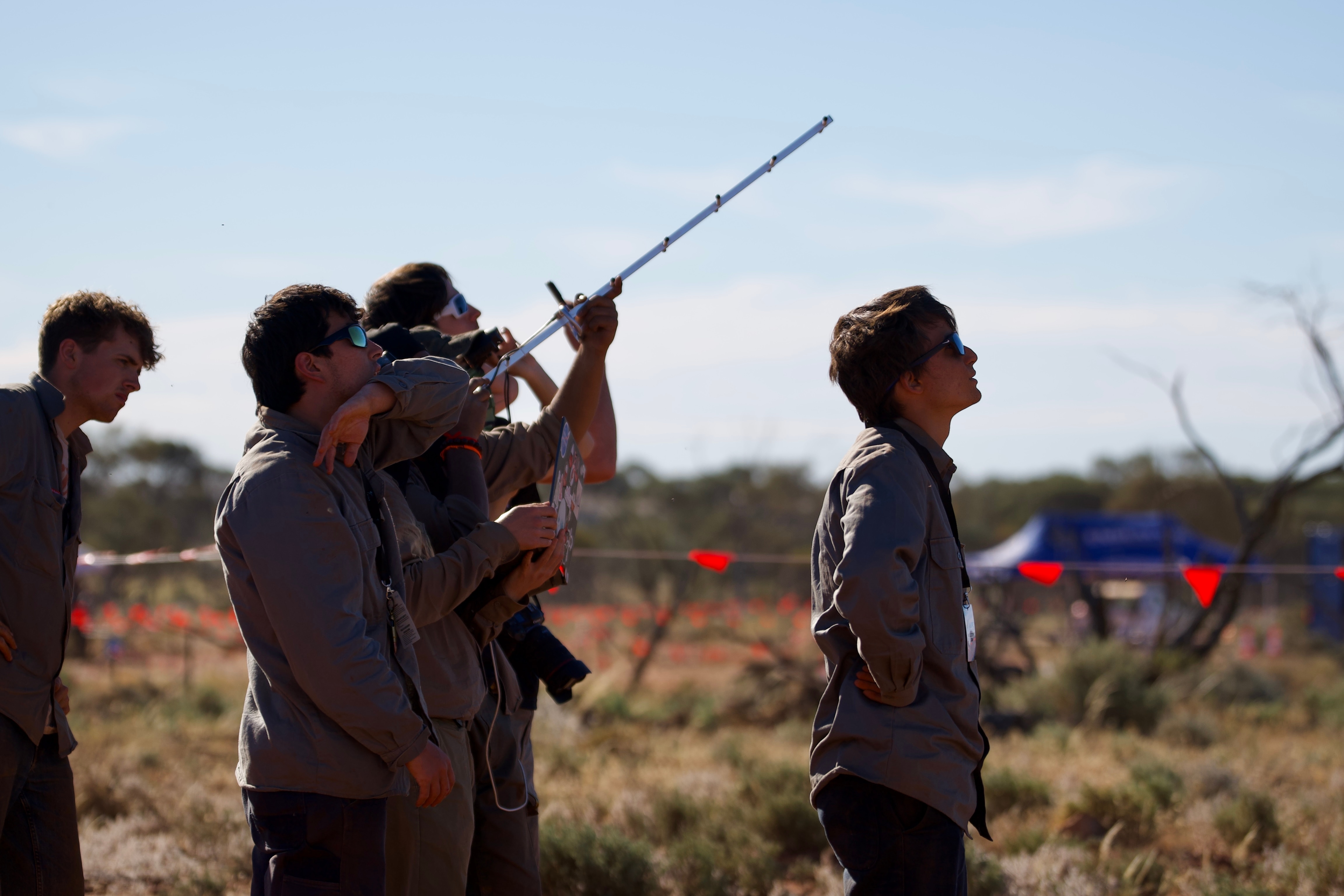 A group of young men in mathcing long-sleeved shirts look to the sky while standing on an outback property.