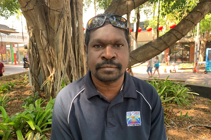 Indigenous man wearing dark shirt with sunglasses on head looks at camera without smiling