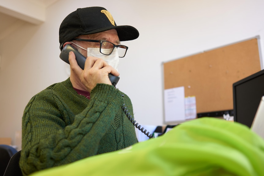 A support worker in a cap and green sweater sits at a computer, talking on the phone.