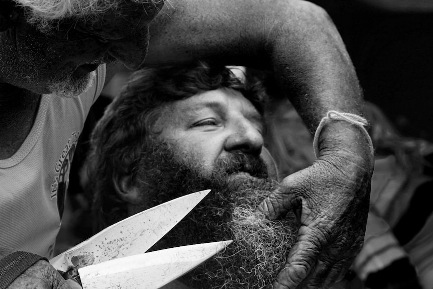 Black and white photo of a man holding large silver scissors to Luke Rowbottom's beard.
