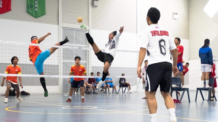 Three men kick a small round ball over a volleyball net.