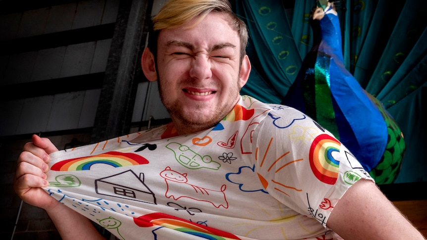 A young man smiles while tugging on a rainbow shirt.