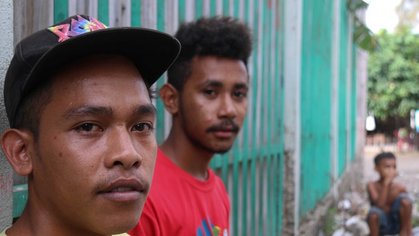 Two young men sit against an aqua and white fence wearing yellow and red shirts.