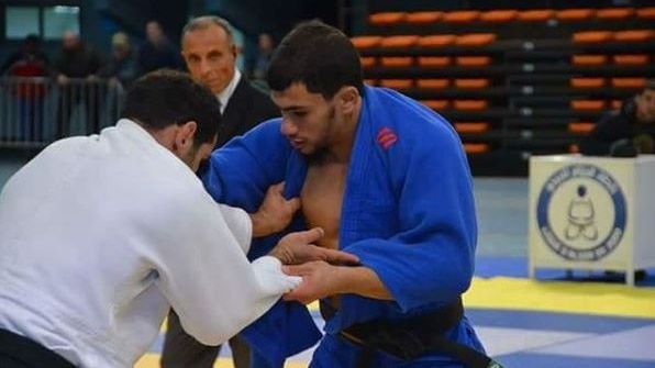 A judoka in a blue outfit grapples with an opponent in a judo competition. 