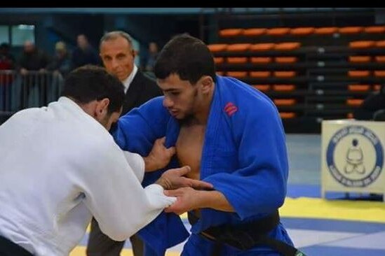 A judoka in a blue outfit grapples with an opponent in a judo competition. 