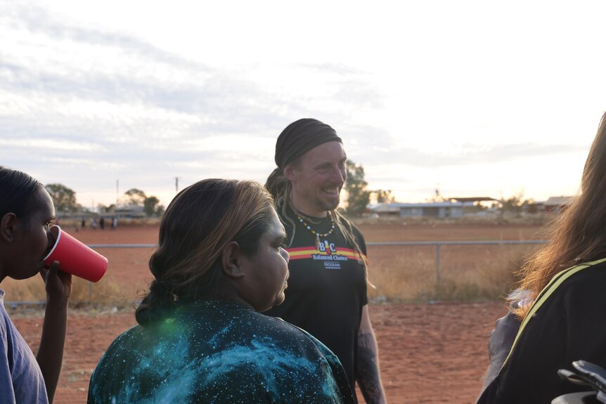 Adam — a young white man — and young First Nations women stand in front of a red dirt oval. Adam is in conversation and smiling.