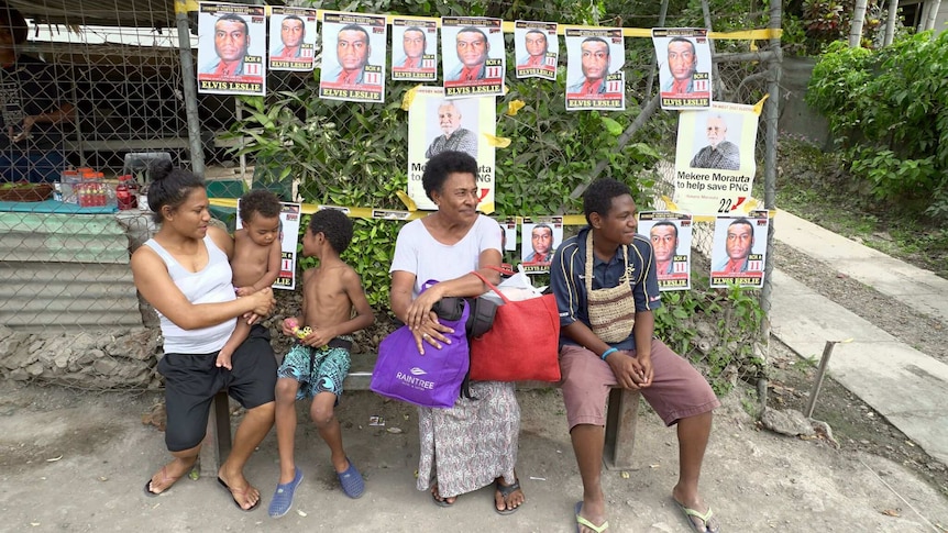 People wait outside a shop decorated with small campaign posters showing two of the candidates' faces in Port Moresby.