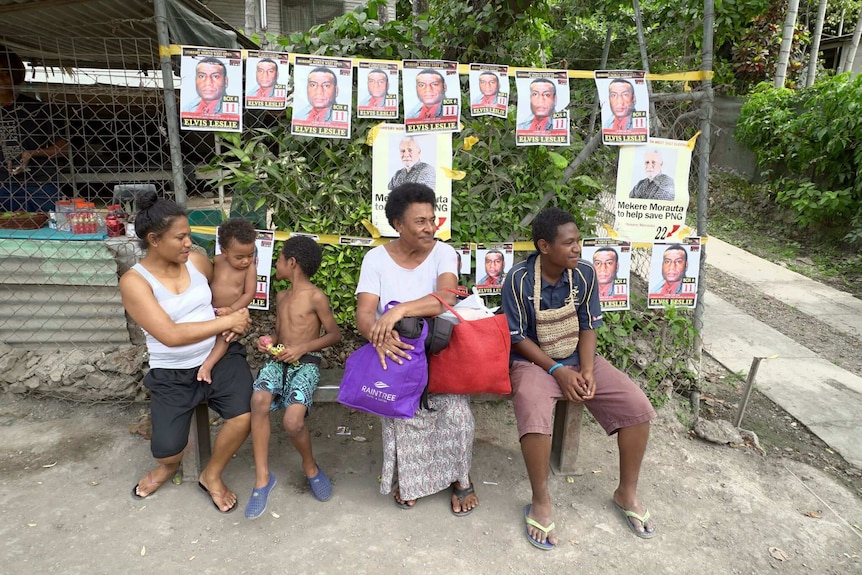 People wait outside a shop decorated with small campaign posters showing two of the candidates' faces in Port Moresby.