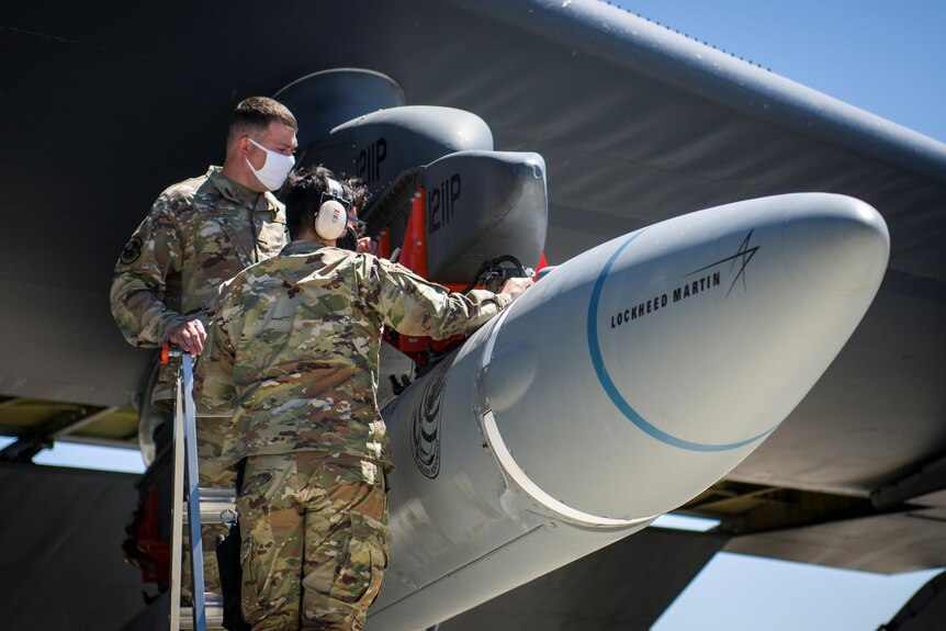 两名身穿军装的男子正在研究安装在飞机机翼下方的导弹。 