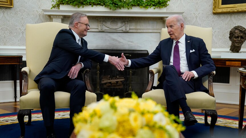 Anthony Albanese and Joe Biden, seated on chairs, shake hands in the Oval Office.