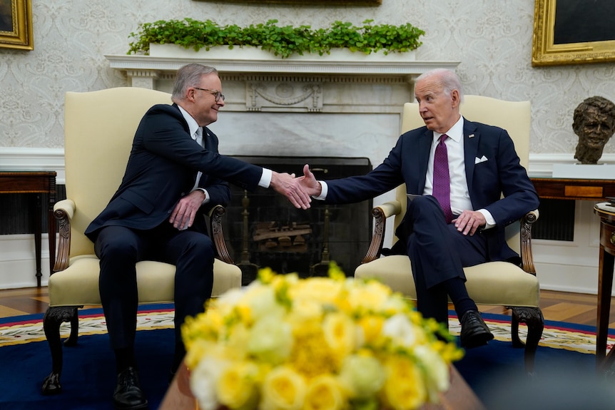 Anthony Albanese and Joe Biden, seated on chairs, shake hands in the Oval Office.