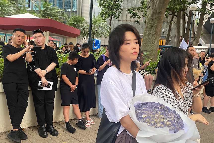 A tearful woman in a white shirt holding a large wreath of purple flowers on a Hong Kong street 
