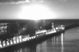 A black and white image shows a large hemisphere of light igniting the horizon over a pier and the bay.