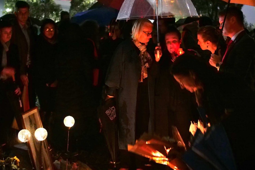 A group of people holding candles in the dark near bunches of flowers.