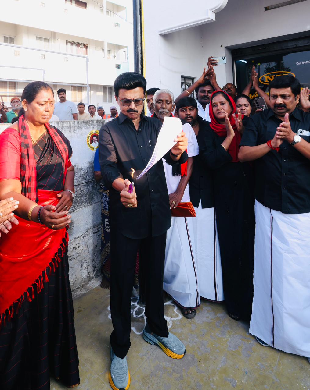 A crowd of people surround a man dressed in black lighting a piece of paper on fire