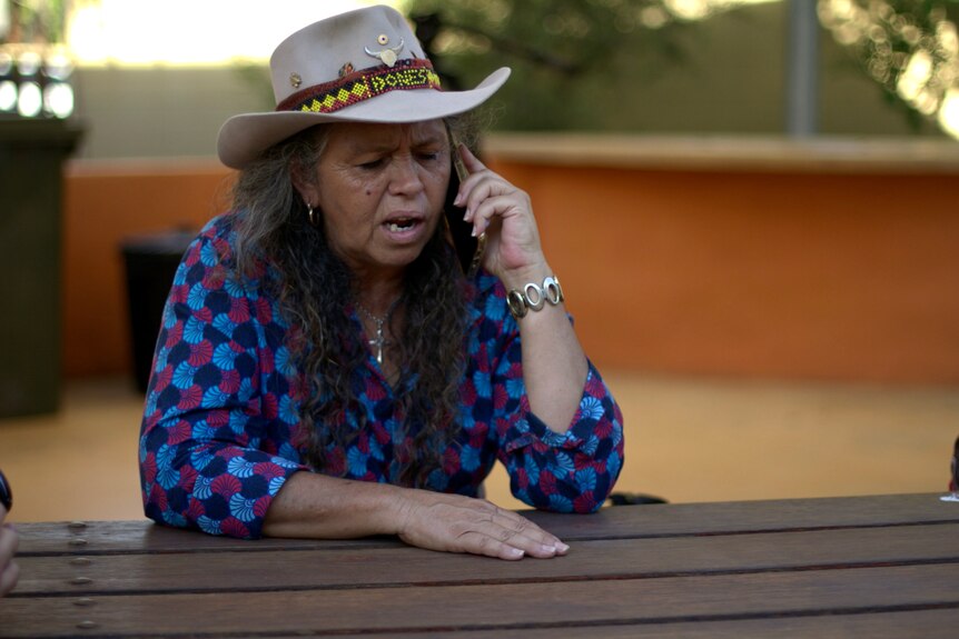 A woman wearing a hat and a checkered shirt makes a phone call while sitting at an outdoor table.