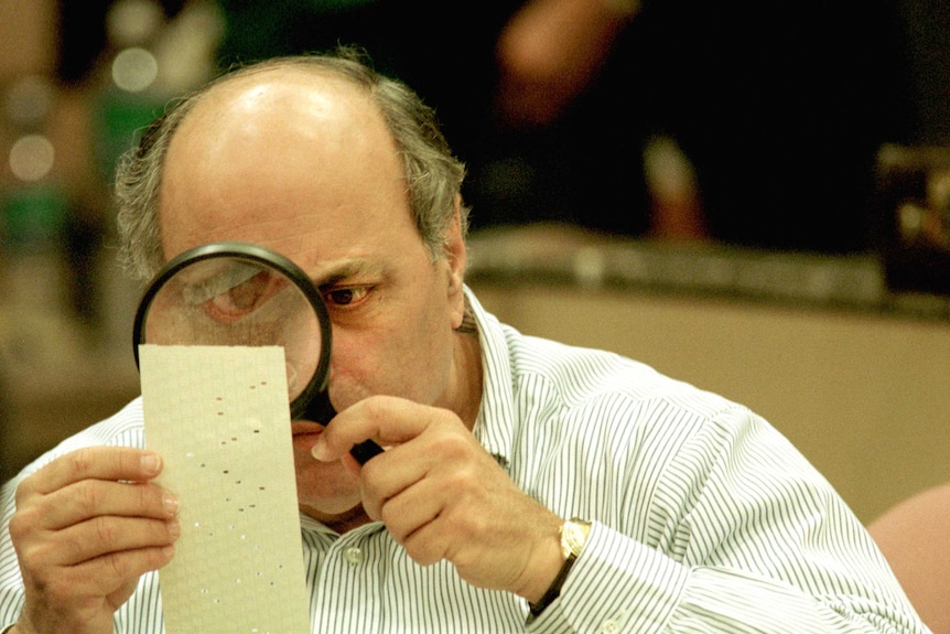 A man examines a punch card with small holes in it by holding a magnifying glass to his eye.
