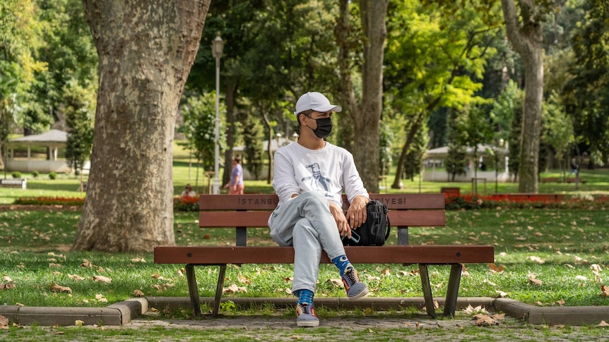 A man in a black face mask and white cap sits at a park bench 