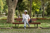 A man in a black face mask and white cap sits at a park bench 