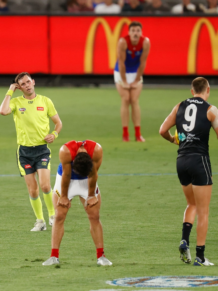 An AFL player bends over with his hands on his hips as an umpire signals behind him during a game. 