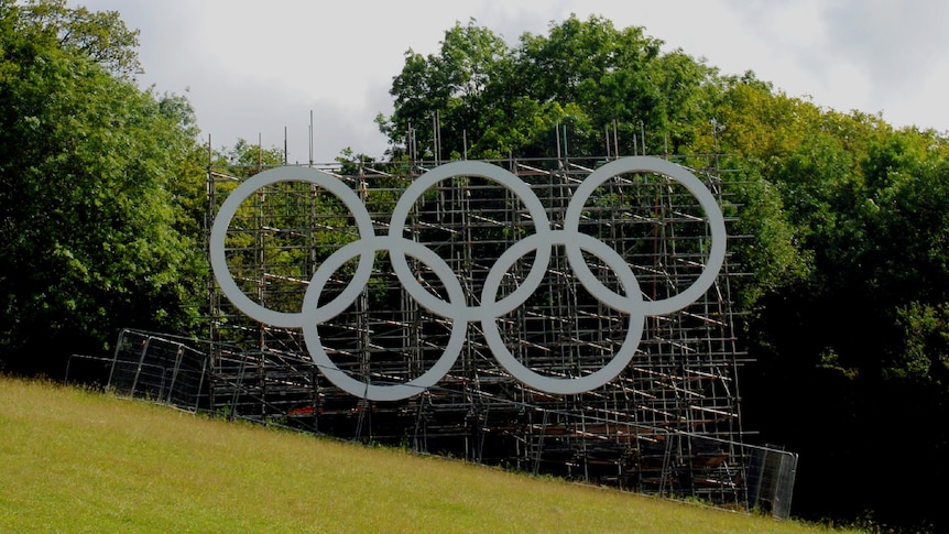 On an overcast day,you view large white Olympic rings mounted on scaffolding that stands perpendicular to a rolling green hill.