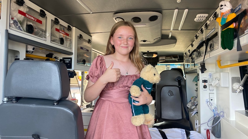 A young girl stands in the back of an ambulance holding a teddy bear