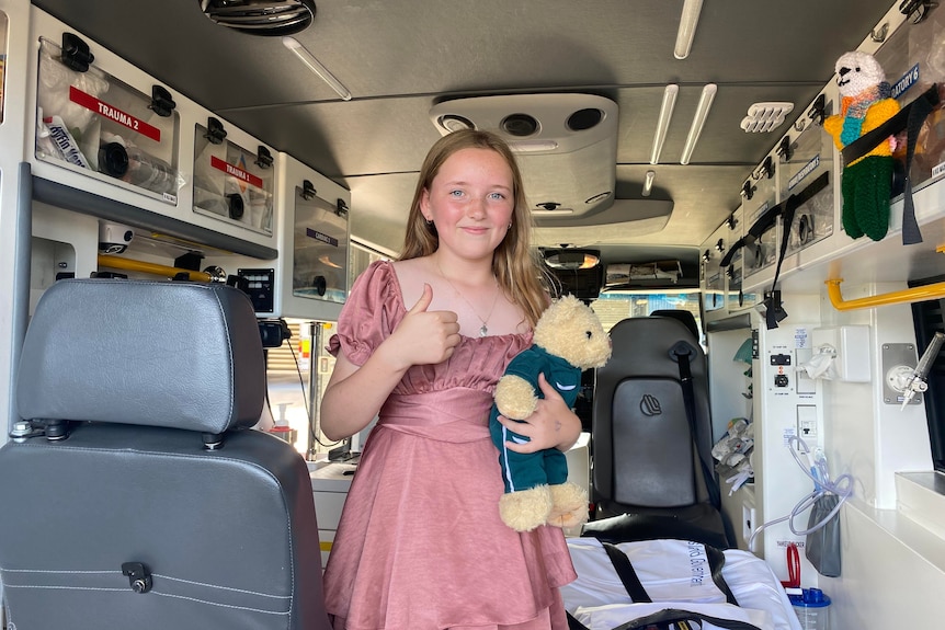 A young girl stands in the back of an ambulance holding a teddy bear