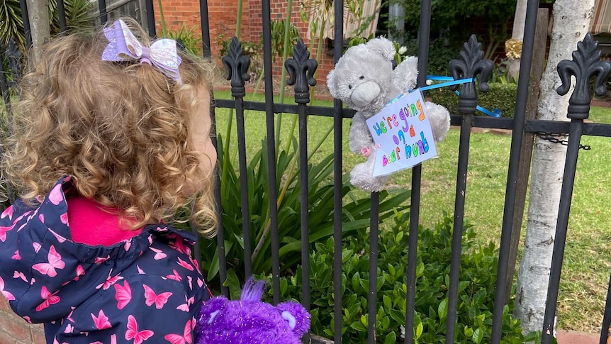 A young girl in a jacket with butterflies on it looks at a bear tied to a fence.
