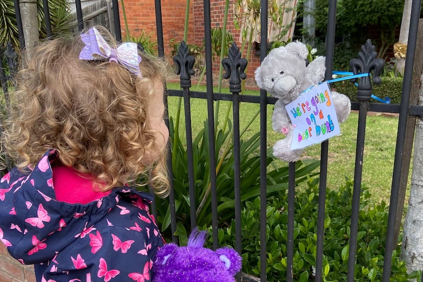 A young girl in a jacket with butterflies on it looks at a bear tied to a fence.