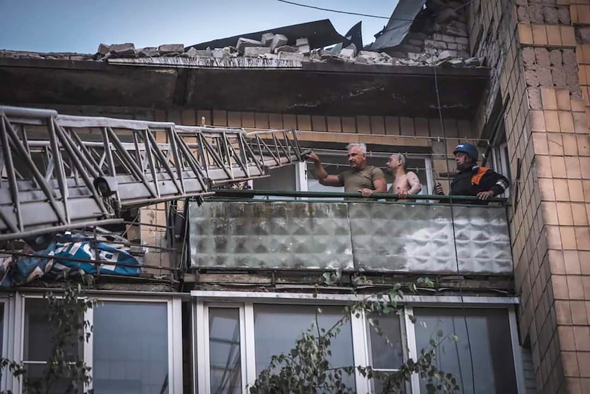 O scară este adusă la balconul unei clădiri distruse, iar trei bărbați o țin în mână 