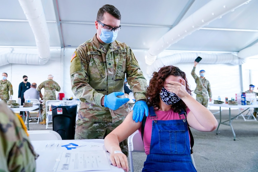 A woman covers her face while a soldier injects a needle into her shoulder