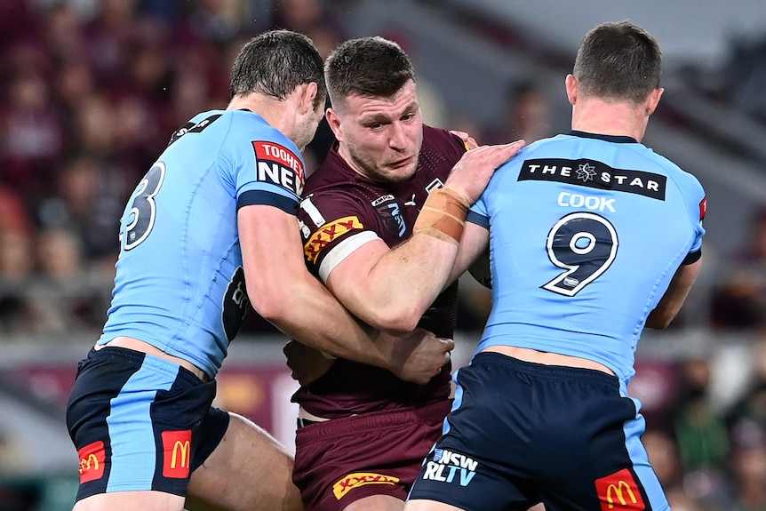 A Queensland State of Origin forward grimaces as he struggles to break the tackles of two Blues defenders.
