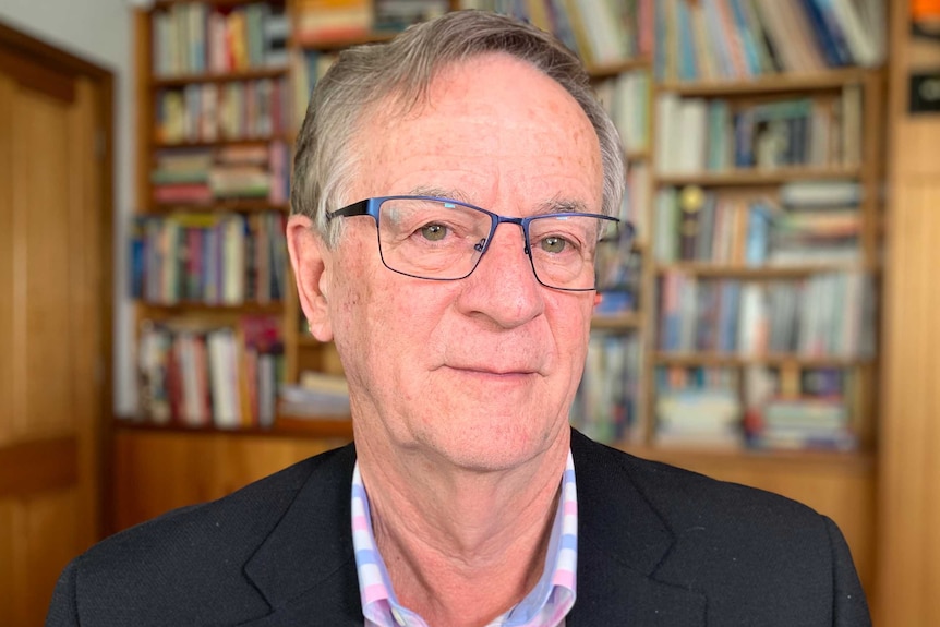 An older, bespectacled fellow wearing a dark blazer sits in front of a bookshelf.