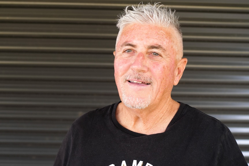 Man with grey hair and grey goatee smiling in front of dark corrugated iron wall.