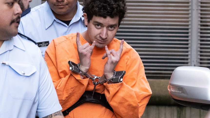 Mert Ney, dressed in orange jumpsuit and in handcuffs gestures with his hands