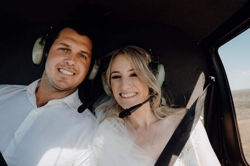 A young man and woman dressed in white, wearing headphones, smile as they sit inside a helicopter.