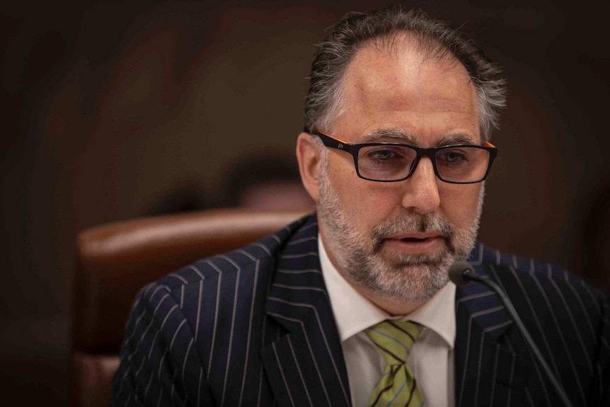 A man wearing glasses and a pinstripe suit sits in a leather chair.