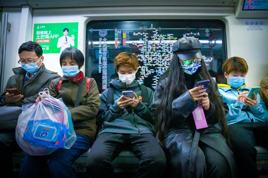 A row of commuters sitting on a train in Beijing, wearing face masks and looking at their phones