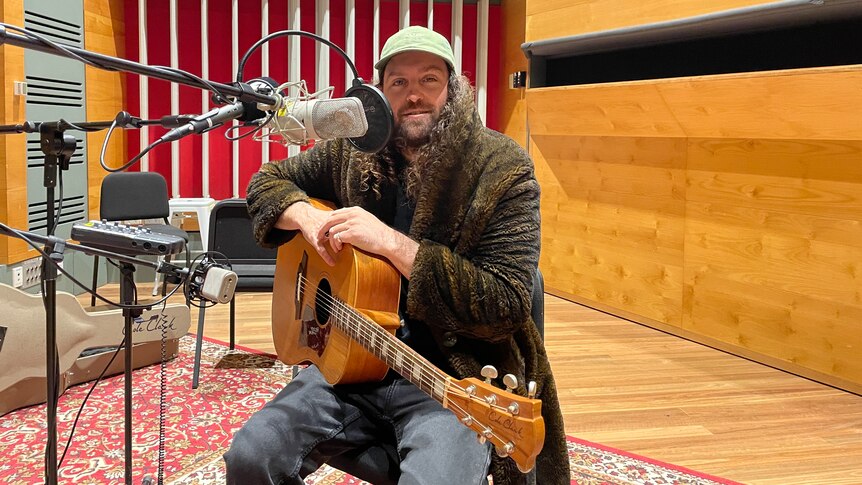 Boo Seeka sits in the ABC recording studio on a stool with a guitar in his lap, cap on, and he is wearing a fur coat