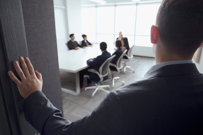 A man can be seen looking into a meeting room from behind. People are sitting around a desk. 