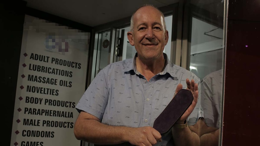 An older man stands outside a sex store holding a rubber paddle.