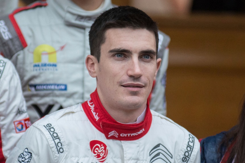 A man with short cropped brown hair in a racing suit.