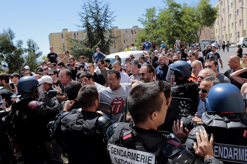 A large crowd of people yelling and being held back by police in Bastia's Lupino neighbourhood, Corsica.