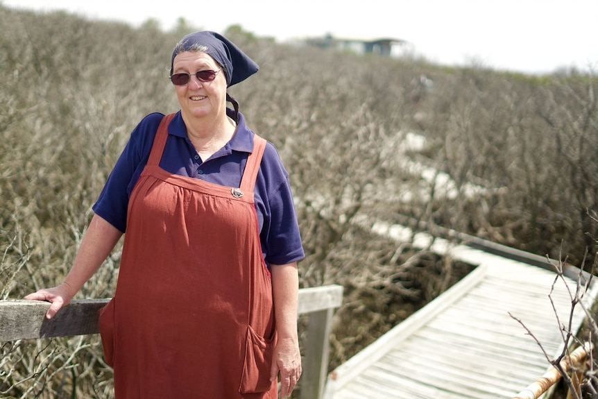 A woman wearing overalls and a bandana leans on a walkway rail surrounded by dead mangroves