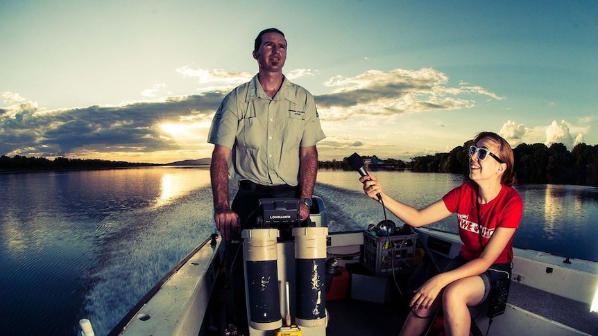 Kaitlyn Sawrey (right) sits in a motorboat holding a microphone up towards a man standing at the boat's controls.