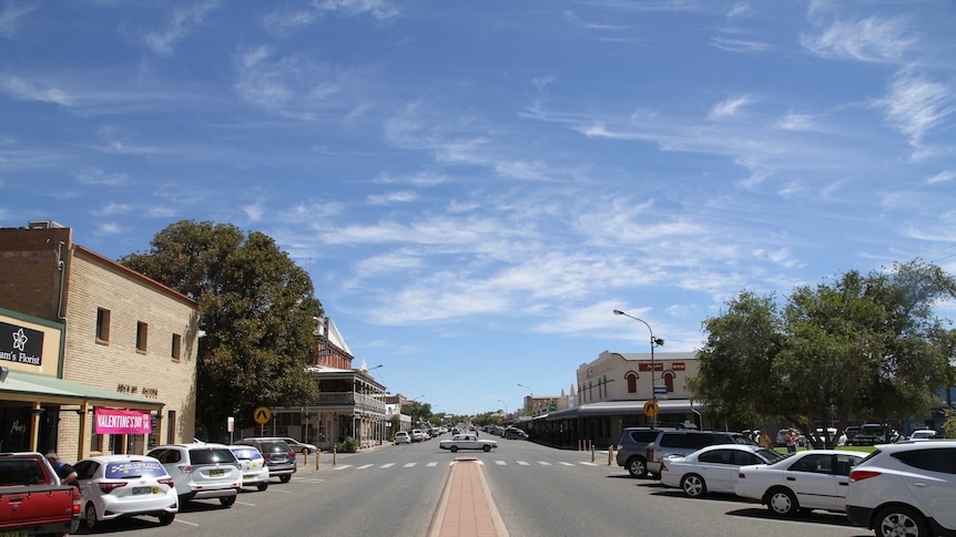 A blue almost cloudless sky with a road in the foreground, cars parked to the side. Symmetrical streetscape