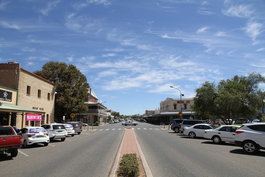 A blue a,lmost cloudless sky with a main, divided street in the foreground, cars parked on an angle on either side.