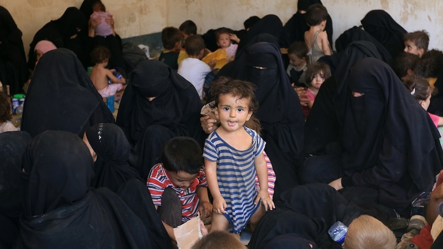 A young girl stands surrounded by women and children in a crowded refugee camp.