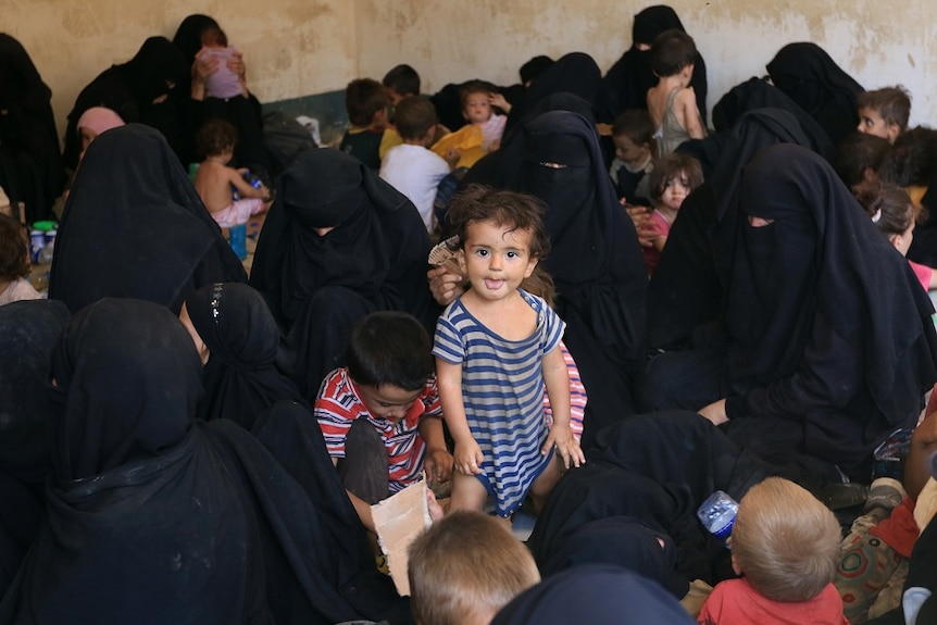A young girl stands surrounded by women and children in a crowded refugee camp.