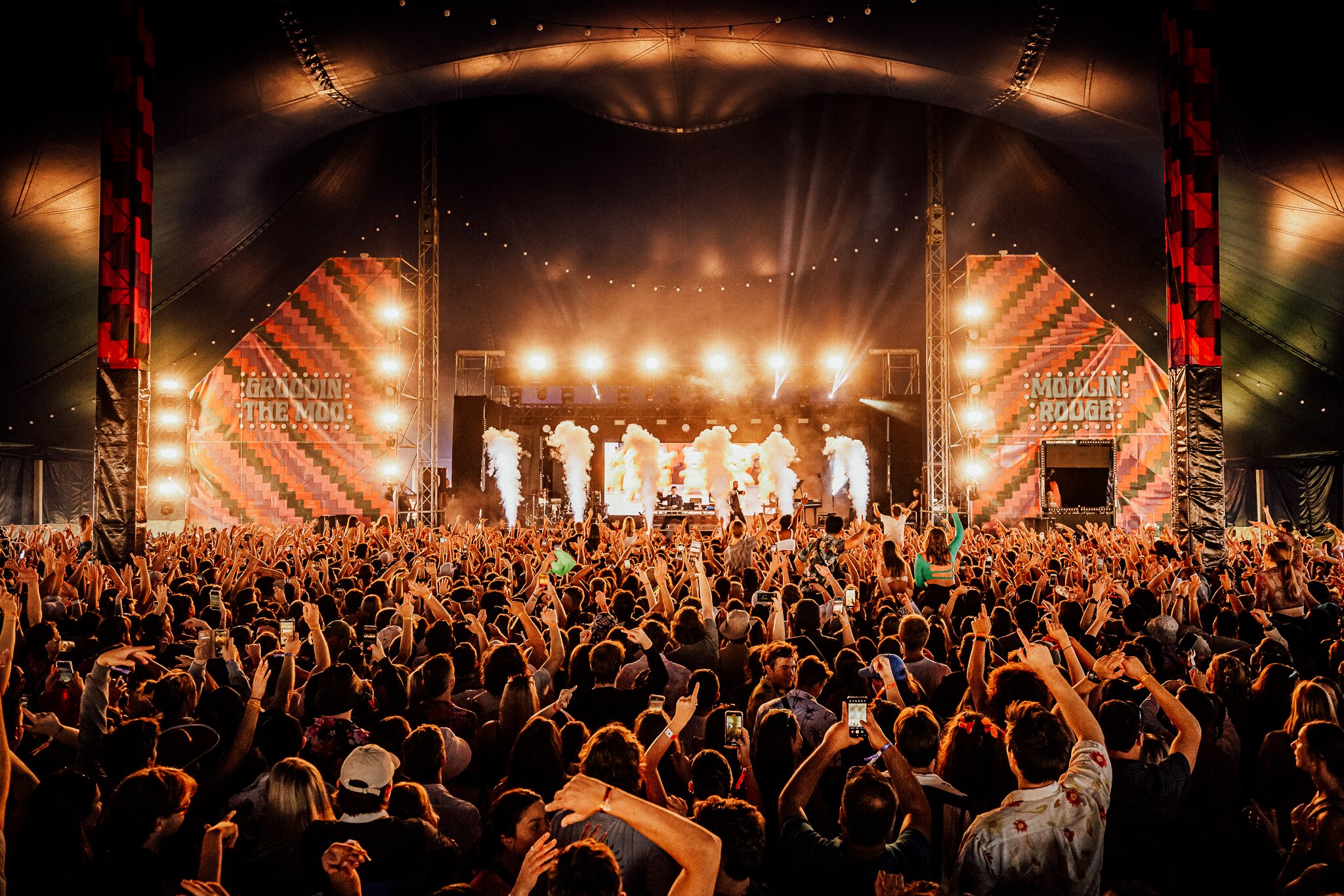 Festival crowd facing a stage with warm white lights underneath a large marquee tent